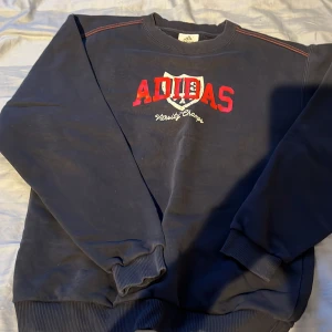 Mörkblå Adidas sweatshirt - Snygg mörkblå sweatshirt från Adidas med rött och vitt tryck på bröstet där det står 'Adidas Varsity Champs'. Tröjan har rund hals, ribbade muddar och klassisk passform. Perfekt för en sportig och avslappnad stil. Passar M,L. 