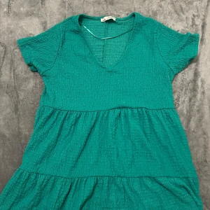 Grön kortklänning från Pull&Bear - Supersöt grön kortklänning från Pull&Bear med v-ringning och korta ärmar. Klänningen har en crinklad struktur och volanger i kjolen som ger en avslappnad och trendig look. Perfekt för varma dagar när du vill vara både bekväm och snygg.