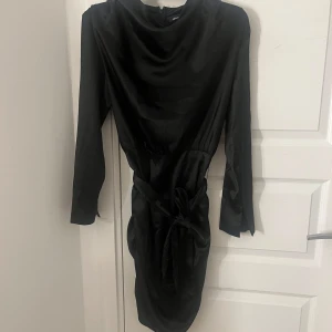 Svart satinklänning Gina Tricot 36 - Supersnygg svart kort satinklänning från Gina Tricot med långa ärmar, knyt i midjan och dragkedja bak. Klänningen har en elegant glansig finish och en figurnära passform som framhäver siluetten samt en slits vid handen. Det är storlek 36 på den och säljs för 150kr