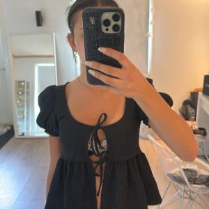 Svart blus med puffärm och knyt - Supersöt svart blus med puffiga korta ärmar och två knyt framtill. Modellen är luftig och har en lätt rynkad struktur i tyget. Djup urringning och flowy passform nedtill ger en trendig vibe. Perfekt att styla med jeans eller kjol.