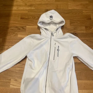 Vit tröja/hoodie från Sail Racing - Vit hoodiejacka från Sail Racing med dragkedja framtill och bröstficka med logga. Jackan har en sportig look, huva och är tillverkad i mjukt fleecematerial. Perfekt för dig som gillar stilren och bekväm streetwear.