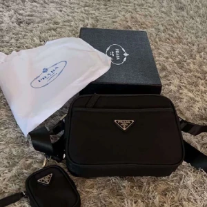 Svart axelväska från Prada - Säljer en svart axelväska från Prada med klassisk trekantslogga i metall framtill. Väskan har justerbar axelrem och en mindre avtagbar ficka. Tillverkad i slitstark nylon med snygga detaljer. Perfekt för dig som vill ha en stilren och lyxig väska.