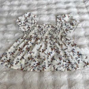 Blommig blus med puffärmar - Supersöt blommig blus i ljusbeige med små bruna och blå blommor. Toppen har smockad överdel, voluminösa puffärmar och en flowy passform nedtill. Perfekt för en romantisk look och passar till både jeans och kjol.