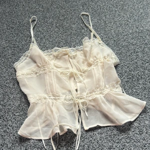 Vit spets korsettopp från Kimchi Blue i XS - Vit korsettopp från Kimchi Blue i storlek XS. Tunt, lätt material med spetsdetaljer, smala axelband och snörning framtill. Det går också att knyta banden runt midjan lösare så blir den lite större. Volangkant nertill och smockad rygg. Fint skick då den aldrig är använd.