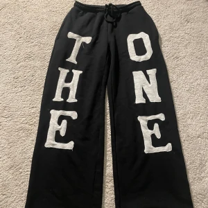 Custom Svarta mjukisbyxor  - Svarta mjukisbyxor med bred passform och stora vita bokstäver som stavar 'THE ONE' på framsidan av benen. Byxorna har resår och snörning i midjan för skön passform. Perfekta för chill dagar eller streetwear-vibe. Custom gjorda 