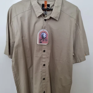 Beige kortärmad skjorta Parajumpers - Stilren beige skjorta från Parajumpers i bomull med korta ärmar och svarta knappar framtill. Klassisk krage och snygga detaljer som orange hängare i nacken. Perfekt för en avslappnad och trendig look.