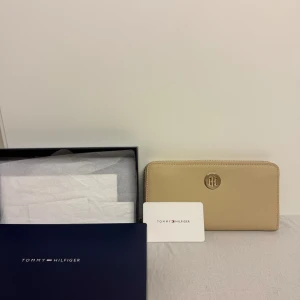 Beige plånbok från Tommy Hilfiger - Snygg beige plånbok från Tommy Hilfiger med guldfärgat TH-emblem framtill. Plånboken har dragkedja runtom och är tillverkad i ett tåligt syntetmaterial. Perfekt för att hålla koll på kort och cash med stil.