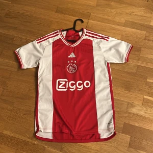 Ajax fotbollströja Adidas röd/vit - Ajax hemmatröja från Adidas med klassiska röda och vita paneler, Ziggo sponsortryck och klubbmärke på bröstet. Tröjan har korta ärmar, rund hals och tre ränder på axlarna. Tillverkad i lätt och ventilerande polyester, perfekt för match eller träning.