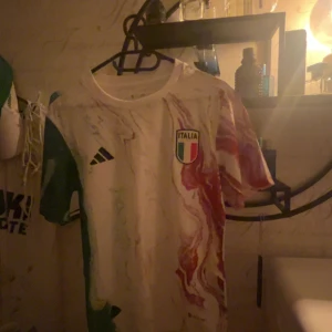 Italien fotbolls tröja - Snygg Italien fotbollströja från Adidas med unikt marmorerat mönster i vitt, grönt och rött. Tröjan har korta ärmar, rund hals och det klassiska Italienska landslagsmärket på bröstet. Perfekt för dig som vill sticka ut på match eller i vardagen.  Pris går att diskutera