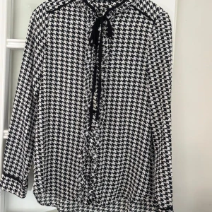 Svartvit blus med knyt från Zara Basic - Snygg svartvit blus från Zara Basic med klassiskt hundtandsmönster. Blusen har långa ärmar, knytband vid halsen och svarta detaljer längs kanterna. Luftigt material som känns skönt mot huden och passar perfekt till jeans eller kjol. XS men passar S också!