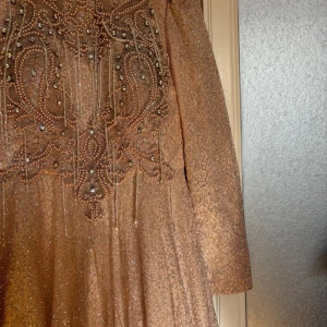 Glittrig långklänning i champagne - Säljer en maxad långklänning i champagnefärg med glittrigt tyg och detaljerad brodyr upptill. Klänningen har långa ärmar och är dekorerad med pärlor och paljetter som ger en lyxig känsla. Perfekt för dig som vill sticka ut med elegans och wow-faktor.