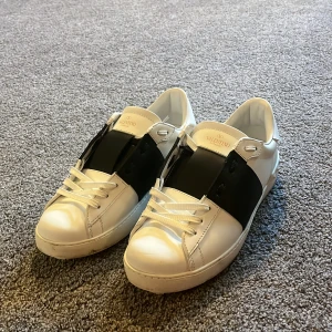 Valentino Garavani Open sneakers vit/svart - Snygga Valentino Garavani Open sneakers i vitt skinn med bred svart läderdetalj över vristen. Klassisk rund tå, vita snören och coola nitar baktill på sulan. Perfekta för dig som gillar stilrena och lyxiga sneakers med edge.