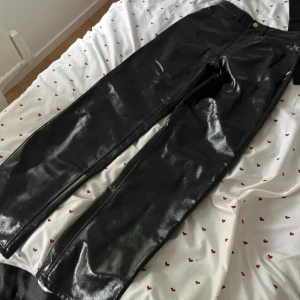 Svarta glansiga byxor i skinnimitation - Säljer ett par svarta glansiga byxor i skinnimitation. De har rak passform och klassisk midja med knapp och dragkedja framtill. Perfekta för dig som vill sticka ut med en edgy look.