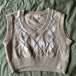 Stickad väst från Pull&Bear - Stickad oversized väst i beige med klassiskt rutigt mönster i vitt och brunt framtill. Djup v-ringning och ribbad kant nedtill samt vid armarna. Perfekt att ha över en skjorta eller t-shirt. Säljer pga att den är för stor för mig! Endast använd en gång.