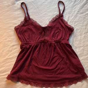 Vinröd linnetopp med spets från Nelly - Supersöt vinröd linnetopp från Nelly med tunna axelband och v-ringning. Toppen har fina spetsdetaljer längs urringning och nederkant samt en liten rosett framtill. 