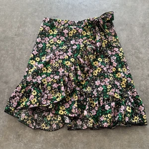 Blommig omlottkjol från Vero Moda XS - Supersöt blommig omlottkjol från Vero Moda i svart med gula, lila och gröna detaljer. Kjolen har volangkant nertill och knyts i sidan för en snygg passform. Perfekt för dig som gillar färg och mönster!
