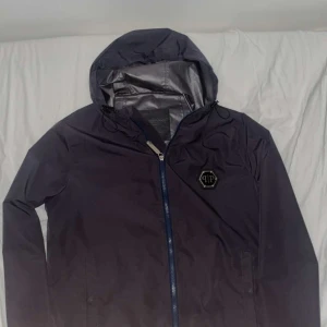 Svart vindjacka från Philipp Plein - Svart vindjacka från Philipp Plein med huva och dragkedja framtill. Jackan har en stilren look med logotyp på bröstet och justerbar dragsko vid huvan. Perfekt för blåsiga dagar och enkel att matcha med olika outfits.