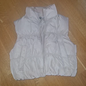 Beige dunväst från H&M Divided XS - Säljer en beige dunväst från H&M Divided i storlek XS. Västen har hög krage, två fickor framtill och dragkedja hela vägen. Den är quiltad och har en puffig, trendig look som passar perfekt till lager-på-lager-stil.