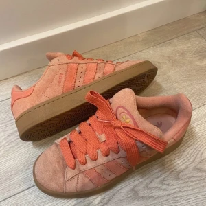 Adidas Campus rosa sneakers i mocka - Snygga Adidas Campus sneakers i en ljusrosa och orange färg med breda matchande skosnören. Skorna har klassiska tre ränder på sidan, gummisula och är tillverkade i mjuk mocka. Perfekta för dig som vill sticka ut med färg och stil. Dom är i bra skick och kan behövas tvättas. 