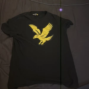 Svart t-shirt med gul örntryck - Svart t-shirt från Lyle & Scott med stort gult örntryck på bröstet. Klassisk passform och rund halsringning. Perfekt för dig som vill sticka ut med en cool och sportig vibe.