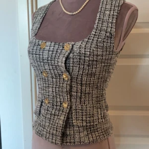 Rutig väst med guldknappar - Snygg rutig väst i svart och beige med markerad midja och sex guldiga knappar framtill. Västen har breda axelband, fyrkantig ringning och är tillverkad i ett strukturerat tyg som ger en exklusiv känsla. Perfekt för att lyfta vilken outfit som helst.