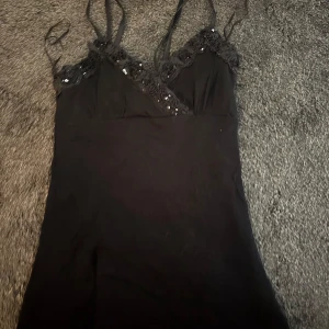 Svart linne med paljettdetaljer - Svart linne med smala axelband och v-ringad omlott framtill. Dekorerad med blommiga spetsdetaljer och paljetter längs urringningen för extra glans. Perfekt för dig som gillar en stilren men ändå festlig look.