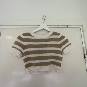 Randig croppad stickad topp - Superfin croppad stickad topp med breda beige och vita ränder. Kortärmad modell med ribbade kanter vid ärmslut, halsringning och nederkant. Perfekt för dig som gillar en chill och trendig stil.