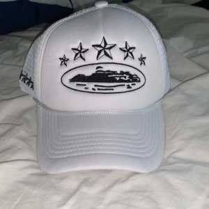 Vit Corteiz keps - Snygg vit keps från Corteiz med svart broderad logga och stjärnor framtill. Kepsen har mesh på sidorna och bak för extra ventilation samt justerbar snapback. Perfekt för dig som vill sticka ut med en stilren och modern accessoar.