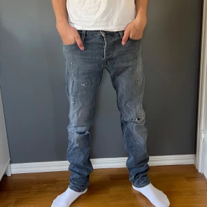 Grå slitna jeans Jack & Jones  - Säljer ett par grå jeans från Jack & Jones med slitna detaljer. Modellen har normal passform och raka ben, klassisk femficksdesign och läderpatch bak i midjan. Perfekta för en avslappnad stil. Storlek 31/34