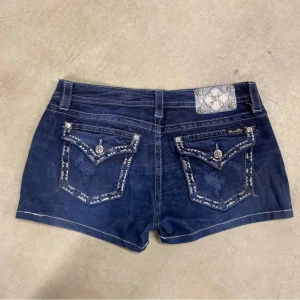 Mörkblå jeansshorts från Miss Me - Dem sitter som en storlek 28!!!!!Snygga mörkblå jeansshorts från Miss Me med dekorativa bakfickor och silvriga detaljer. Shortsens fickor har mönstrad kant och coola knappar. Klassisk femficksmodell med låg midja och kontrastsömmar. Perfekta för sommaren och en avslappnad stil.