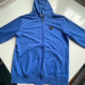 Comme des Garçons play zip hoodie - Blå zip hoodie från Comme des Garçons Play i storlek L. Tröjan har dragkedja, huva och Ikoniskt svart hjärta med ögon på bröstet och stort tryck på ryggen. Tillverkad i mjuk bomull, perfekt för en chill och stilren look.