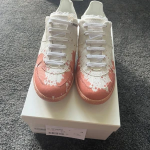 Maison Margiela Replica sneakers färgstänk - Maison Margiela Replica sneakers i vitt med rödbruna färgstänk. Klassisk siluett med rund tå, snörning och platt sula. Ovandel i canvas och detaljer i skinn. Unik design med konstnärlig touch, perfekt för dig som vill sticka ut.