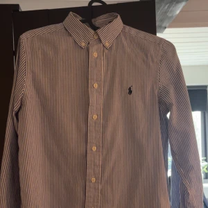 Randig skjorta från Ralph Lauren - Klassisk skjorta från Ralph Lauren med blå och vita ränder, broderad logga på bröstet. Perfekt för sommaren! Skicket är utmärkt då den är nästan helt ny. Hör av dig vid minsta fundering!!
