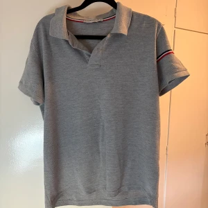 Grå Moncler pikétröja med ränder - Snygg grå pikétröja från Moncler i bomull. Klassisk krage och korta ärmar med coola röd, vit och marinblå ränder på ena ärmen. Perfekt för dig som gillar stilrena och sportiga vibes. Passar till jeans eller shorts för en clean look.