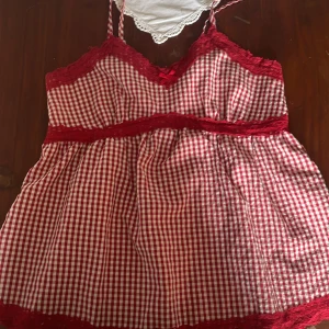 Rutig röd och vit linnetopp med spets - Supersöt linnetopp i rödvitt rutigt mönster med tunna axelband. Toppen har röda spetsdetaljer längs byst, midja och nederkant samt en liten rosett framtill. Dragkedja i sidan för enkel påklädning. Köpt i England! Passar en mindre s till xs.