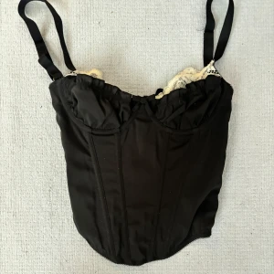 Svart korsett med spetsdetaljer - Snygg svart korsett med justerbara axelband och vita spetsdetaljer upptill. Toppen har markerade sömmar framtill som ger en cool siluett och en feminin touch med spetsen. Perfekt att styla med jeans för en trendig look.