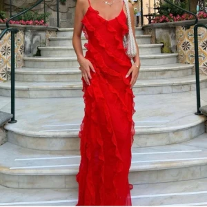 Röd långklänning med volanger - Snygg röd långklänning med tunna axelband och volangdetaljer över hela klänningen. Klänningen har en slits på sidan och en figurnära passform som ger en elegant siluett. Perfekt för dig som vill sticka ut med en färgstark look.