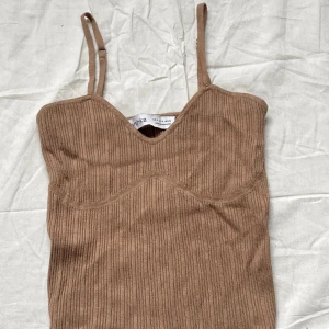 Ribbad brun linnetopp från Bershka - Supersnygg ribbad brun linnetopp från Bershka med smala justerbara axelband och v-ringad skärning. Toppen har en figurnära passform och är perfekt att styla med jeans eller kjol. Materialet är mjukt och stretchigt för en bekväm känsla. Längden är aningen kortare än hellängd, men inte croptop-kort. 