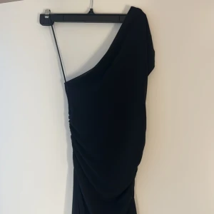 Svart oneshoulder långklänning - Snygg svart långklänning med oneshoulder-design och draperad effekt längs sidan. Klänningen har en elegant och stilren look, perfekt för dig som vill sticka ut. Materialet känns mjukt och följsamt mot kroppen.