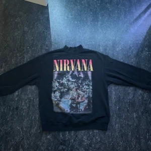 Svart Nirvana sweatshirt Unplugged - Svart sweatshirt med stort Nirvana-tryck fram och motiv från Unplugged in New York. Baksidan har låtlista i gult och vitt. Tröjan har rund hals, långa ärmar och en relaxed passform. Perfekt för dig som älskar bandmerch och vill sticka ut.