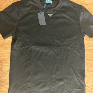 Svart t-shirt från Prada med logga - Prada T-shirt i svart. Stilren, bekväm och i ny skick. INTE ANVÄND. Storlek S.
