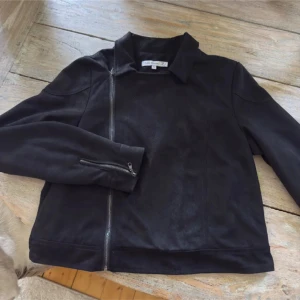 Svart bikerjacka från Flash, stl 38 - Svart bikerinspirerad jacka från Flash i mjuk polyester med sned dragkedja och klassisk krage. Jackan har långa ärmar med dragkedjedetaljer och en clean, stilren look. Perfekt för dig som gillar en edgy vibe och vill ha något som funkar till många outfits.
