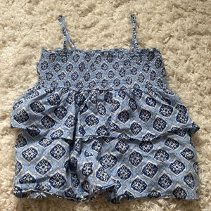 Blå mönstrad linnetopp Gina Tricot - Supersöt blå och vit linnetopp från Gina Tricot med smala axelband och smock upptill. Toppen har ett unikt mönster i olika blå nyanser och volangdetaljer som ger en luftig känsla. Perfekt för varma dagar och enkel att matcha med shorts eller kjol.