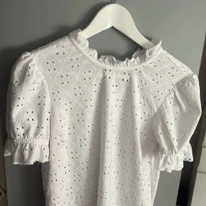 Vit blus med volangkrage från Vila - Supersöt vit blus från Vila med broderade detaljer och hålmönster. Blusen har puffiga korta ärmar med resår och volang vid ärmslut samt en volangkrage som ger en romantisk vibe. Storlek Xs