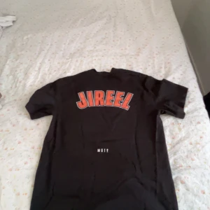 Svart Jireel t-shirt med tryck - Säljs inte längre! Svart t-shirt med stort JIREEL-tryck i rött och vitt på ryggen och ett coolt grafiskt tryck med texten 'LÄR DIG ÄLSKA DIG SJÄLV' på framsidan. Klassisk rund hals och korta ärmar. Perfekt för dig som gillar svensk hiphop och streetwear. 
