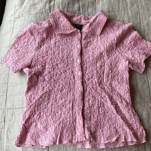 Rosa skrynklig skjorta från Lindex - Supersöt rosa skjorta från Lindex i storlek S. Skjortan har korta ärmar, klassisk krage och knappar framtill. Materialet är skrynkligt och ger en cool textur. Perfekt för dig som vill ha en unik och trendig look.