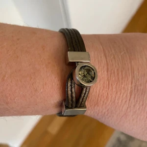 Armband!ä från Dyrberg & Kern - Mycket stilrent och Snyggt brunt armband från Dyrberg/Kern med en stor sten i mitten. Armbandet har en stilren design med flera smala remmar och en metallisk detalj som håller stenen på plats. Perfekt för att ge en elegant touch till din outfit.Längden är 19 cm inkl allt.  Vikt 15 gram.  0,9cm brett 