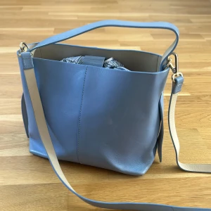 Ljusblå axelväska i skinn från and other stories - Snygg ljusblå axelväska i slätt skinn med stilrena guldfärgade metalldetaljer. Väskan har både axelrem och handtag, samt en rymlig insida med ett randigt innerfoder. Perfekt för dig som vill ha en trendig och praktisk väska med minimalistisk look.