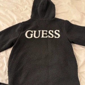 Svart teddyjacka med huva från Guess - Mysig svart teddyjacka från Guess i storlek XS. Jackan har en stor huva och är tillverkad i fluffigt teddy-material. Stor vit GUESS-logga på ryggen ger en clean och trendig look. Perfekt för kalla dagar när du vill vara både varm och snygg.