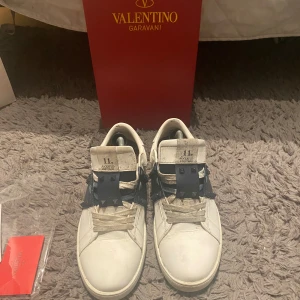 Valentino Garavani Rockstud sneakers - Snygga vita Valentino Garavani Rockstud sneakers med bred marinblå rem över vristen dekorerad med ikoniska nitar. Skorna har rund tå, platt sula och klassisk snörning. Tillverkade i skinn och levereras med originalförpackning och tags. Varan är såklart äktighetsverifierad